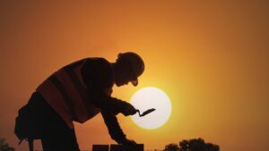 A bricklayer works against a backdrop of orange sky and bright sun, suggesting high temperatures.