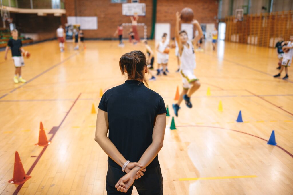 A woman basketball coach watches her team play with her back to the camera.