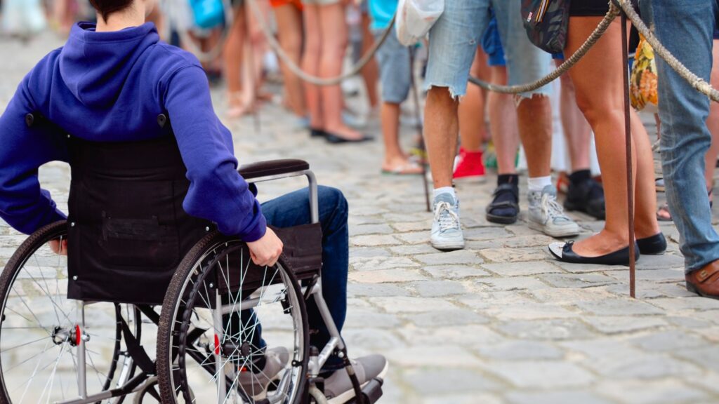 A young man in a wheelchair, seen from the neck down, waits beside a crowded line.